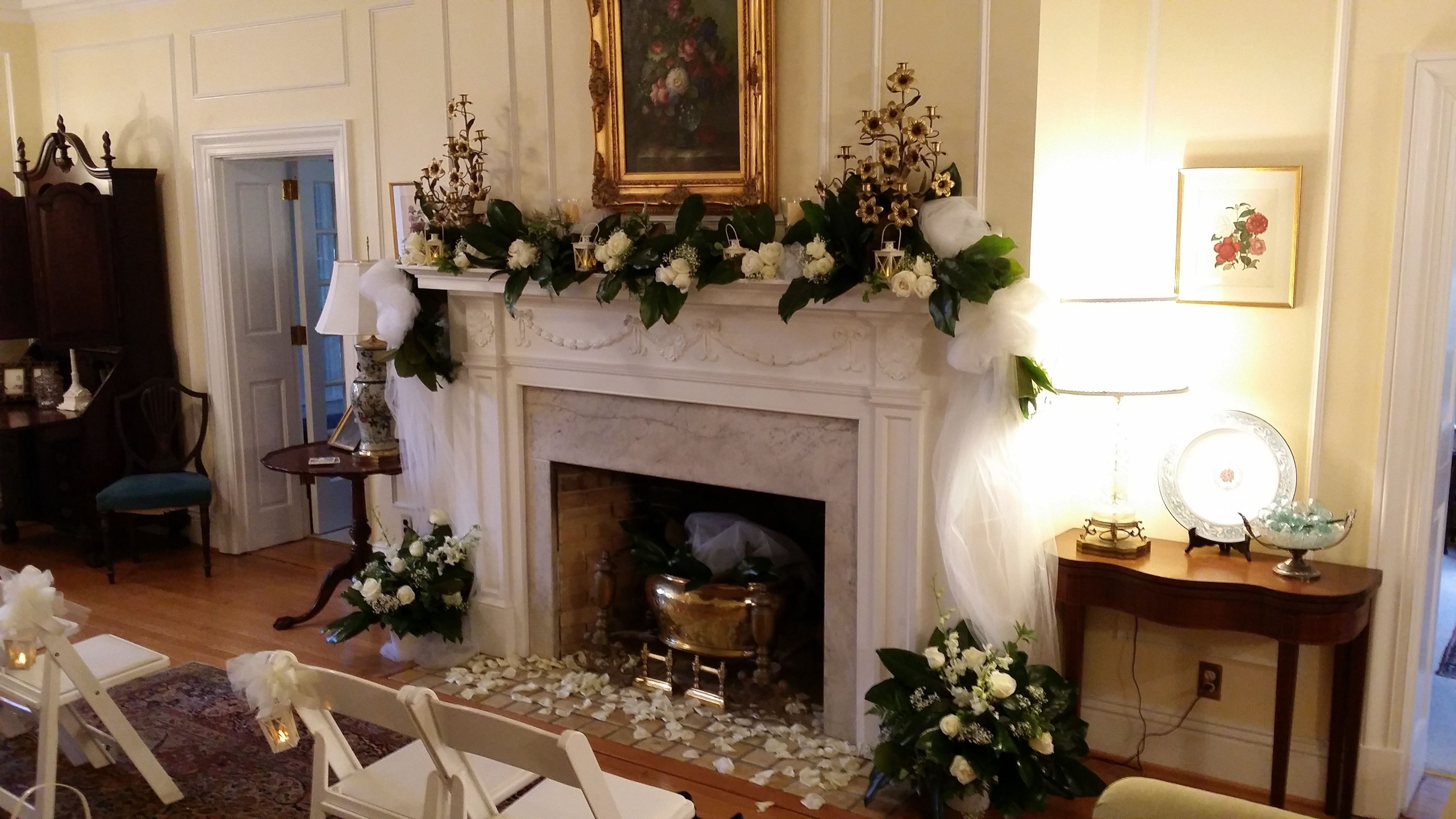 A room with a fireplace, floral arrangements, and a framed painting above it.