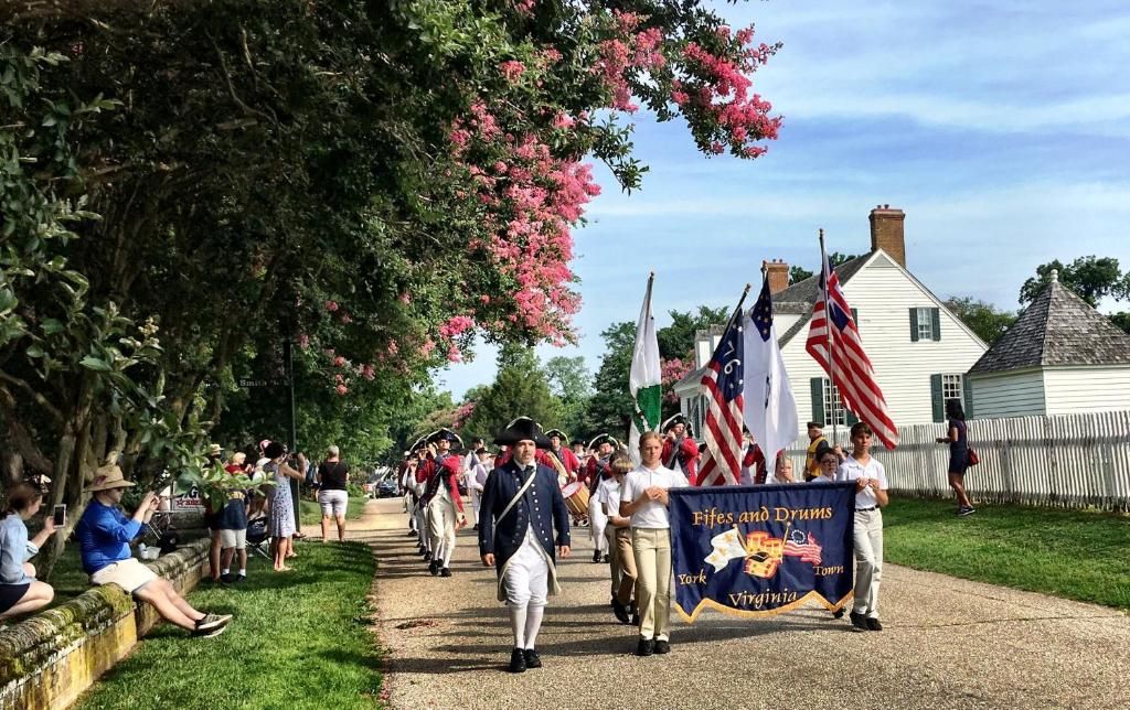 A historical reenactment with participants in period costumes and flags, a pathway lined with spectators, and a building in the background.