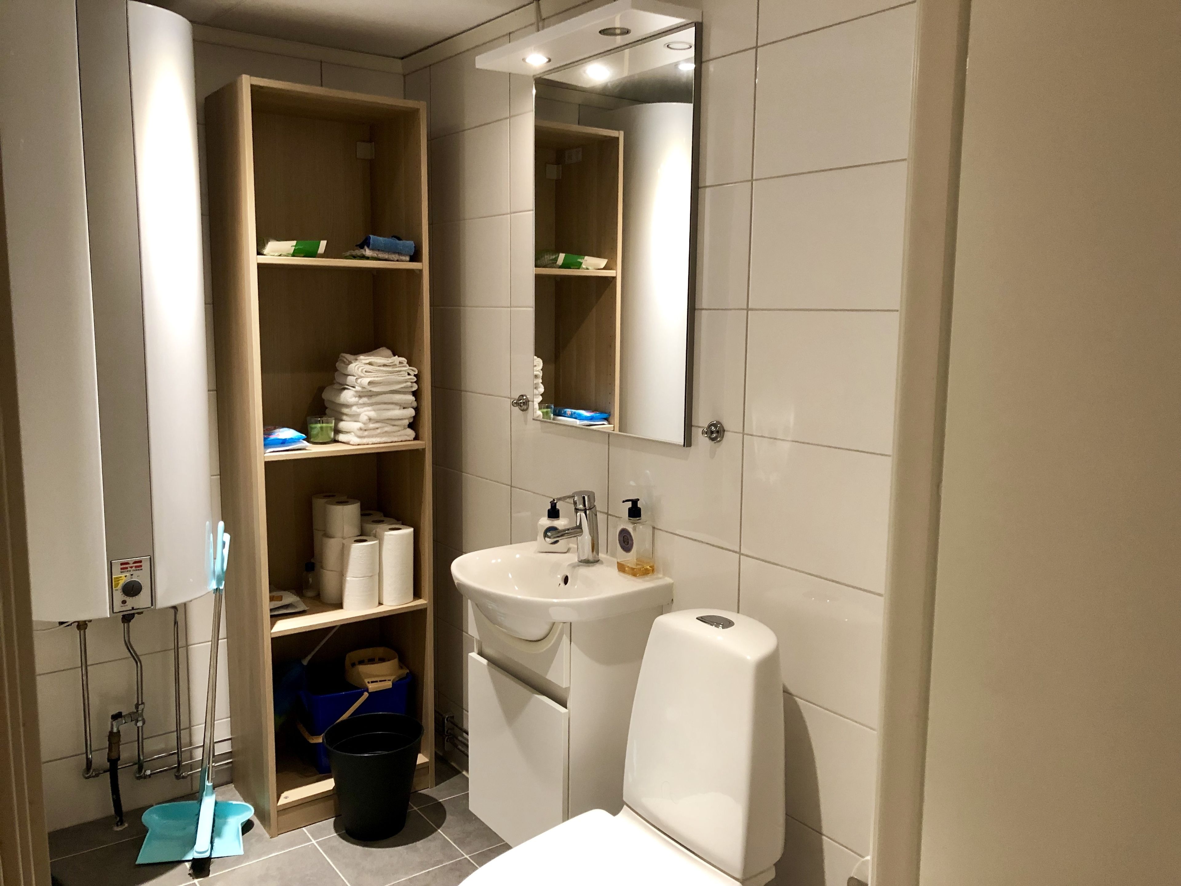 Un baño con lavamanos, espejo, repisas con toallas, papel higiénico y un trapeador.