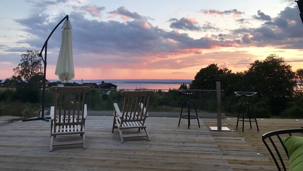 A wooden deck with two wooden chairs and a white umbrella, overlooking a body of water during sunset. - Hills Loft Cottage with Sauna (Gränna)