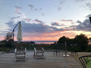 A wooden deck with two wooden chairs and a white umbrella, overlooking a body of water during sunset. - Hills Loft Cottage with Sauna (Gränna)