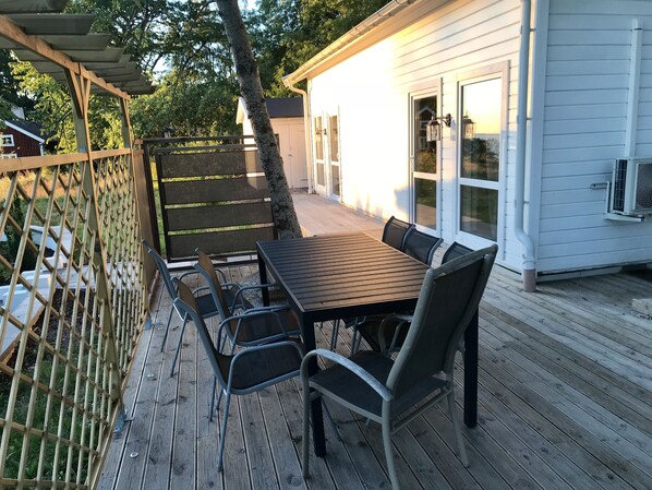 A covered outdoor patio with a wooden table and chairs, a metal fence, and a white building in the background. - Hills Loft Cottage with Sauna (Gränna)