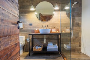 A modern bathroom with a wooden wall, a round mirror, a sink with two faucets, and a glass shower enclosure. - Hotel Desertica (San Pedro de Atacama)