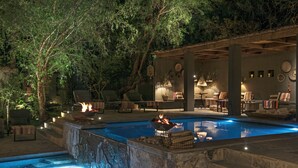 Un espace piscine agrémenté d’un escalier en pierre, d’un foyer extérieur, de coins salon en plein air et d’une terrasse couverte avec des tables et des chaises.