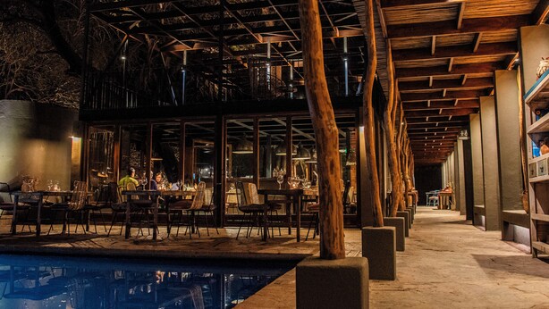 Un espace piscine intérieur aménagé avec des tables et des chaises, des poutres en bois et une structure en verre.