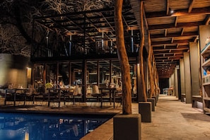 Un espace piscine intérieur aménagé avec des tables et des chaises, des poutres en bois et une structure en verre.