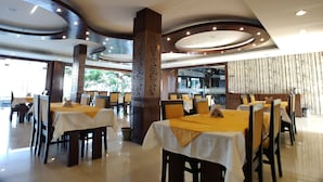 A spacious restaurant with multiple tables and chairs, a central decorative pillar, and a large window providing a view of the outdoors. - Hotel Shiv Villas (Goharganj)