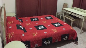 Un dormitorio con una cama cubierta con una colcha roja y motivos blancos y negros, un escritorio de madera, una silla y un espejo.