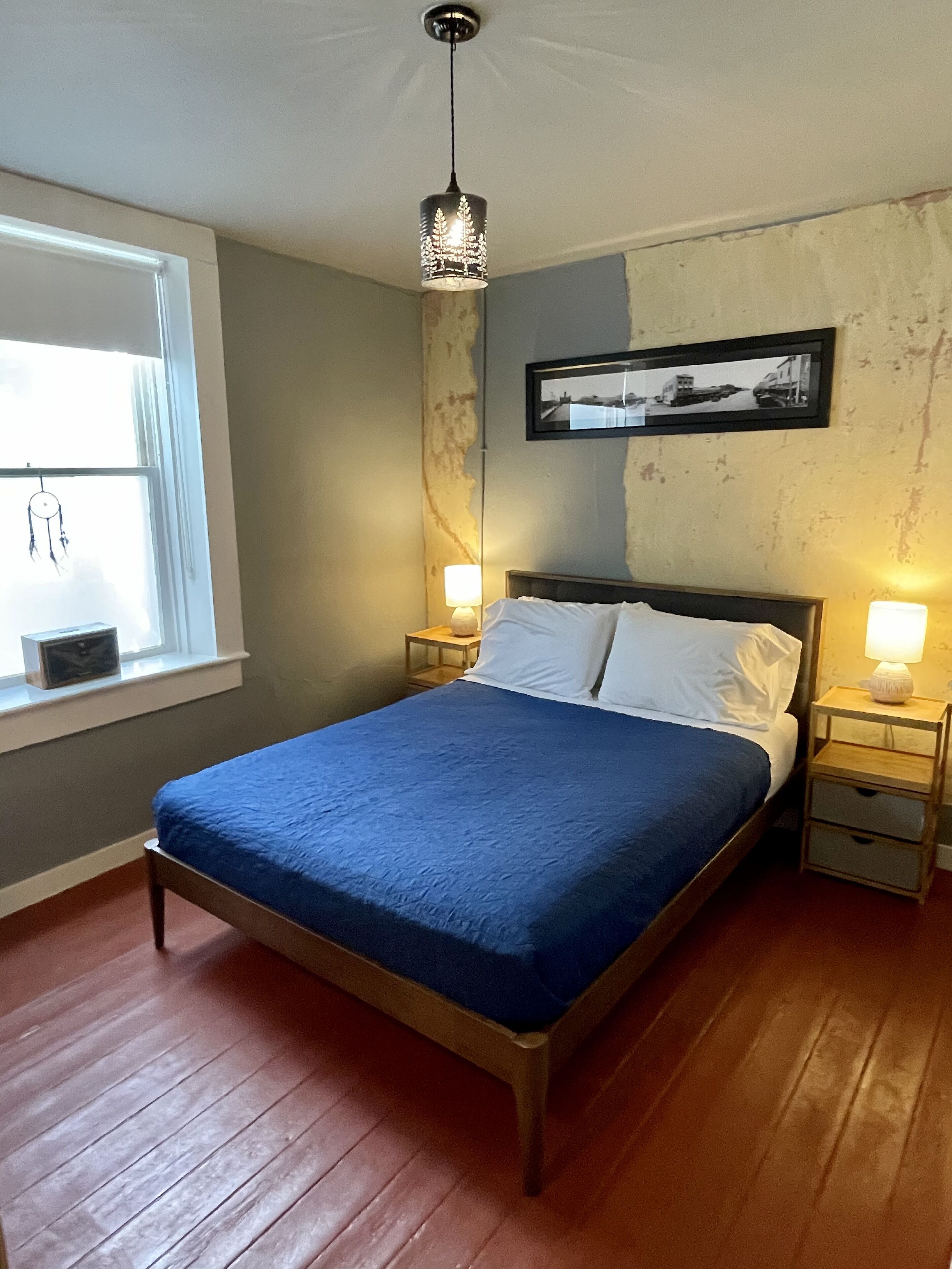 A bedroom with a wooden bed, two bedside tables with lamps, a window with curtains, and a framed picture on the wall.