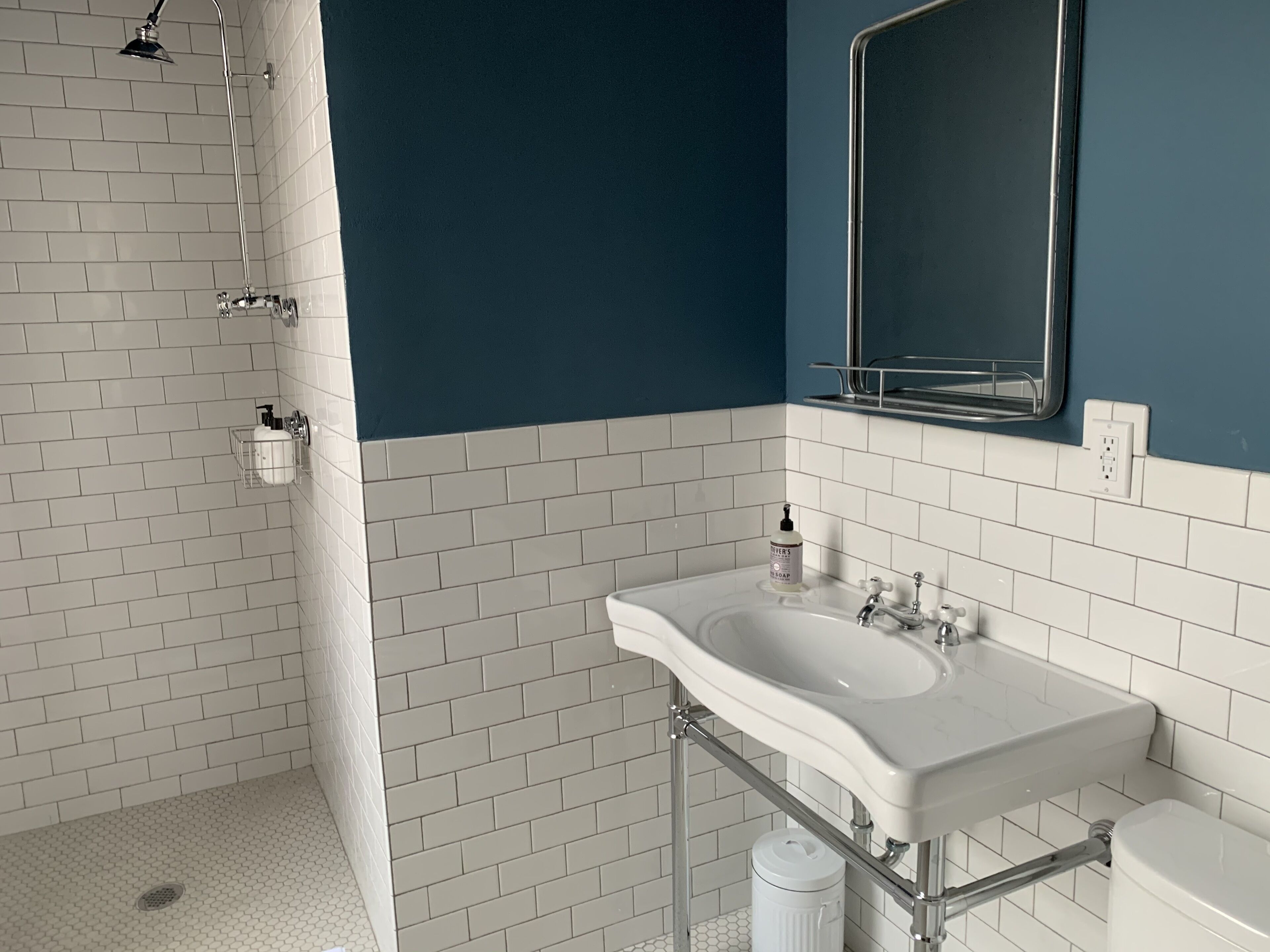 Une salle de bain avec des carreaux de céramique blancs rectangulaires, une douche avec une porte en verre, un lavabo blanc avec un miroir et un mur d’accent bleu.