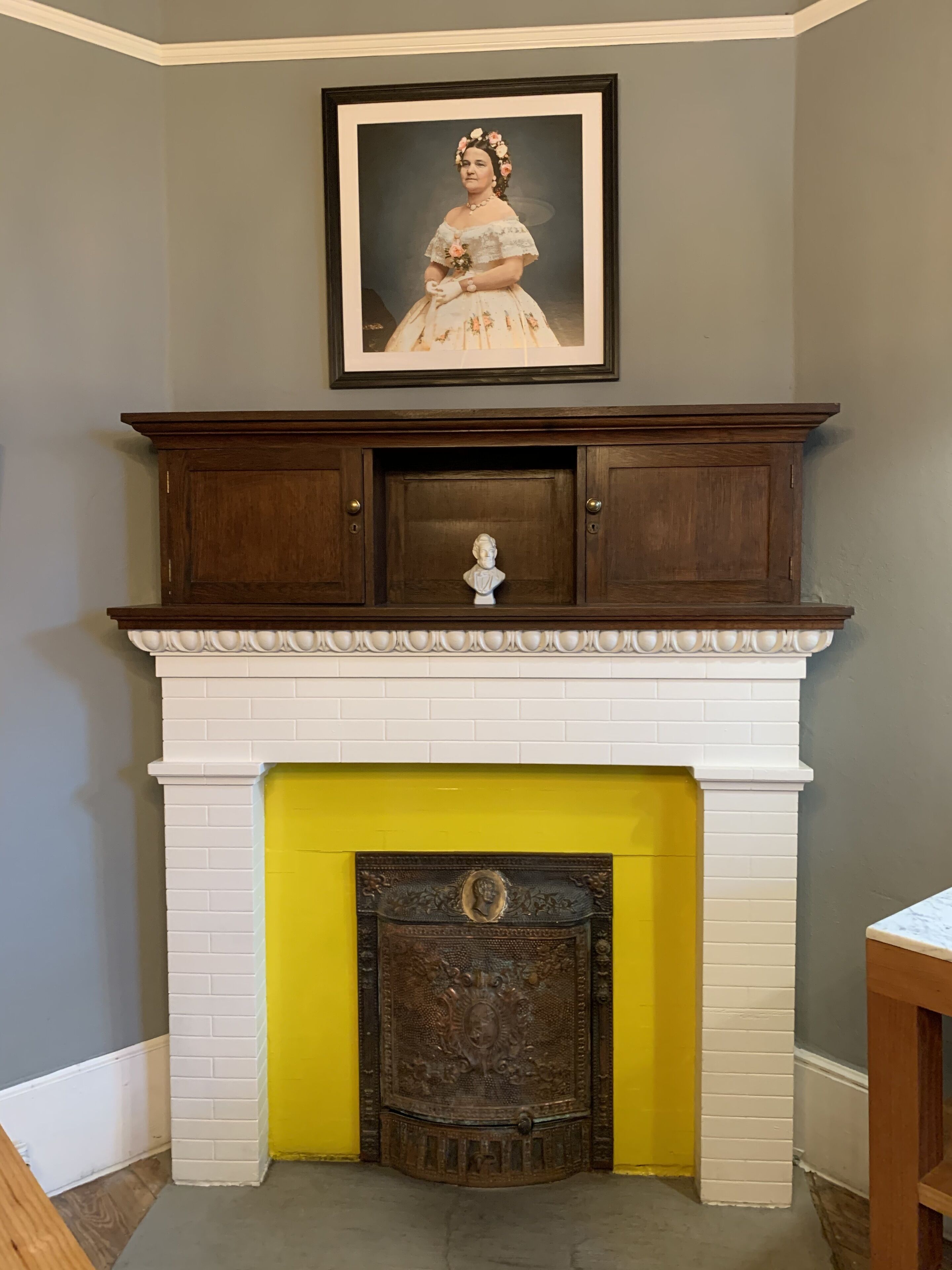 A fireplace with a white brick surround and a yellow accent wall. Above the fireplace, there is a framed portrait.