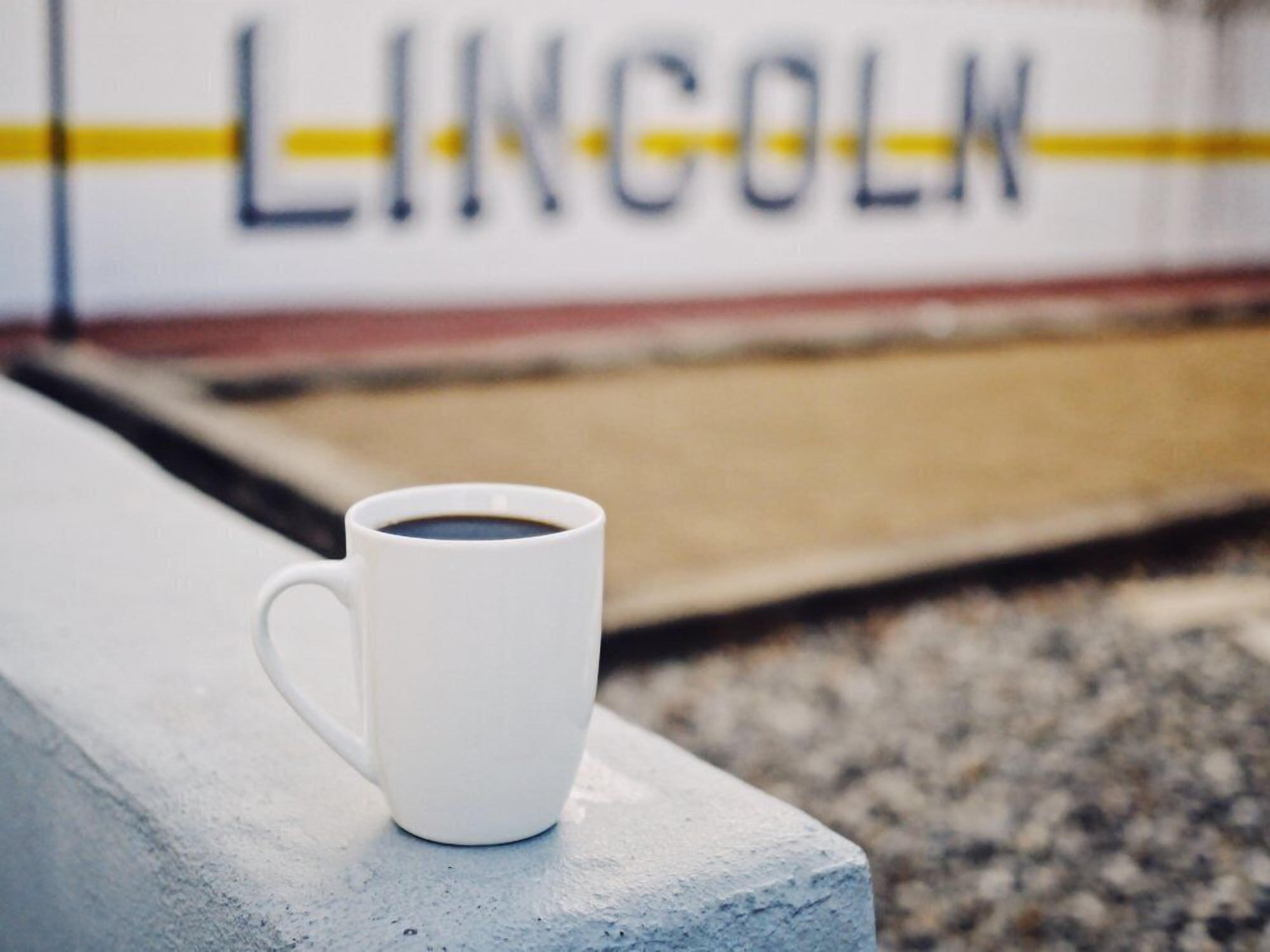 A white mug on a concrete ledge with a blurred background featuring a sign with the word "LINCOLN".