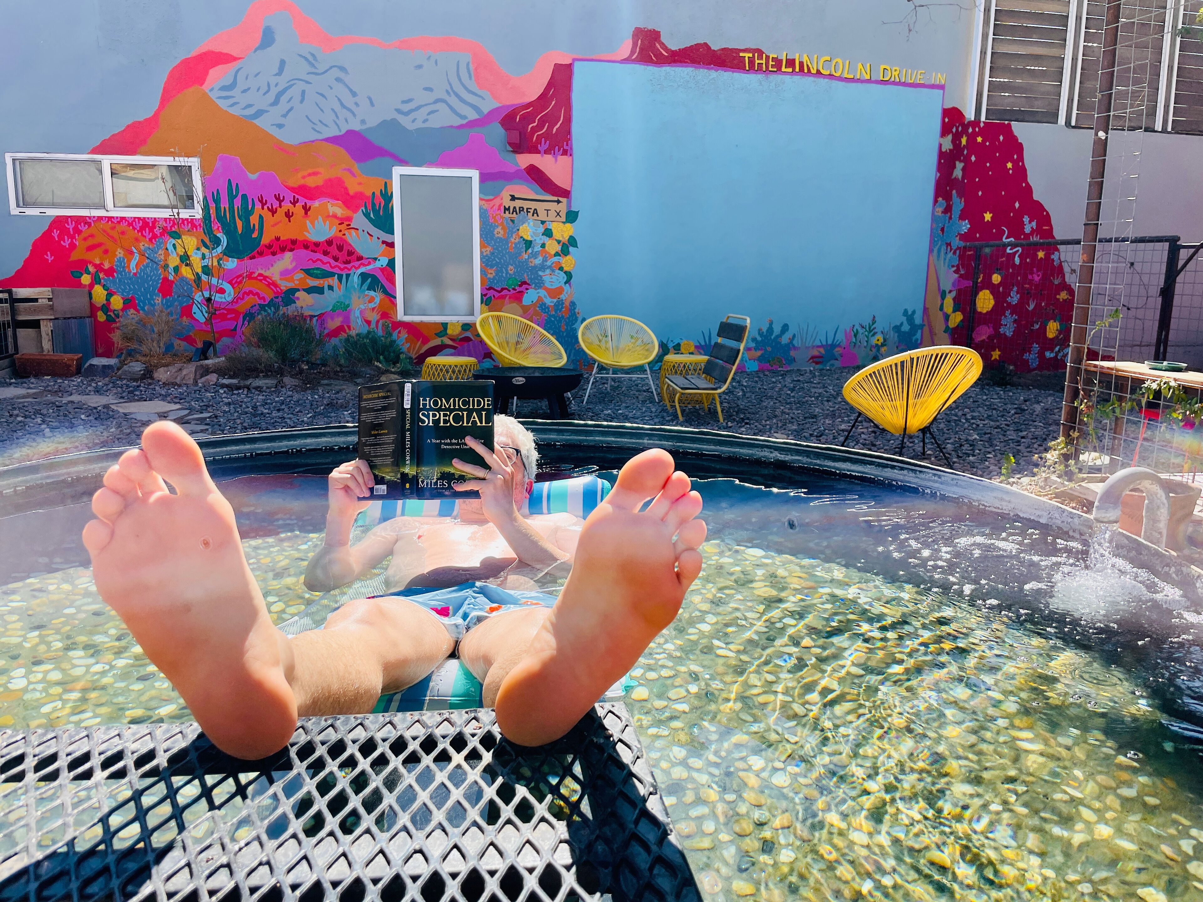 Eine Person entspannt sich in einer Badewanne, im Hintergrund ist ein farbenfrohes Wandbild zu sehen.