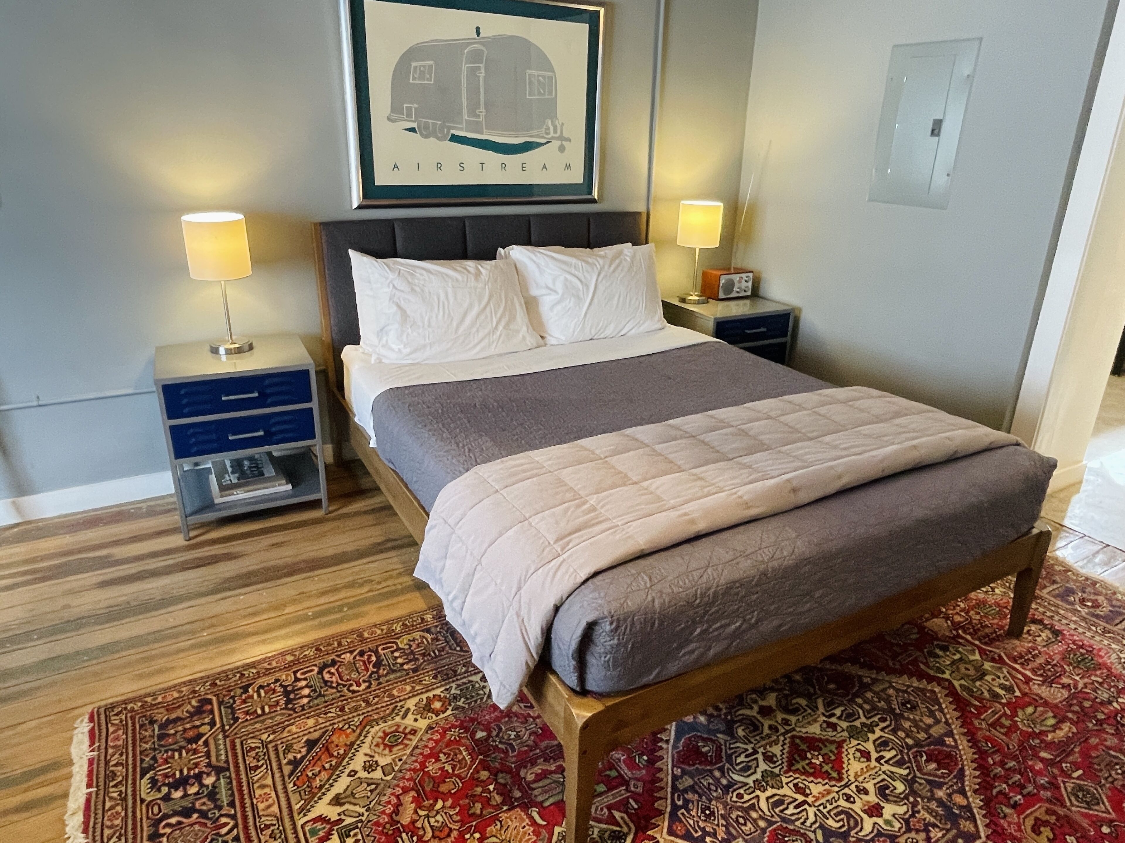 Een net opgemaakt bed met een grijs-beige deken, twee nachtkastjes met lampen, een lijst met een foto van een caravan, en een vloerkleed met een patroon op een houten vloer.