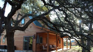 A wooden cabin with a covered porch, large windows, and a view of the surrounding landscape. - Prow'd House Bed & Breakfast (Wimberley)