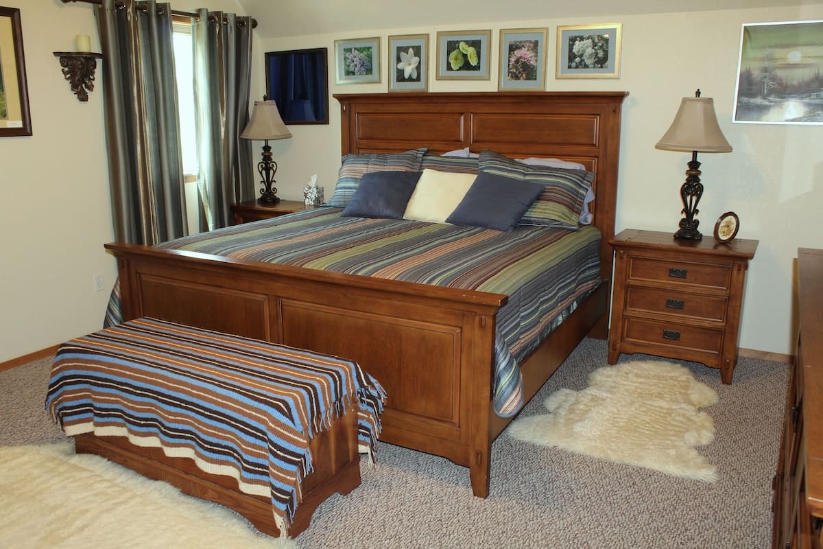 A bedroom with a wooden bed, a bedside table, a nightstand, a lamp, a rug, and a wall with framed pictures.