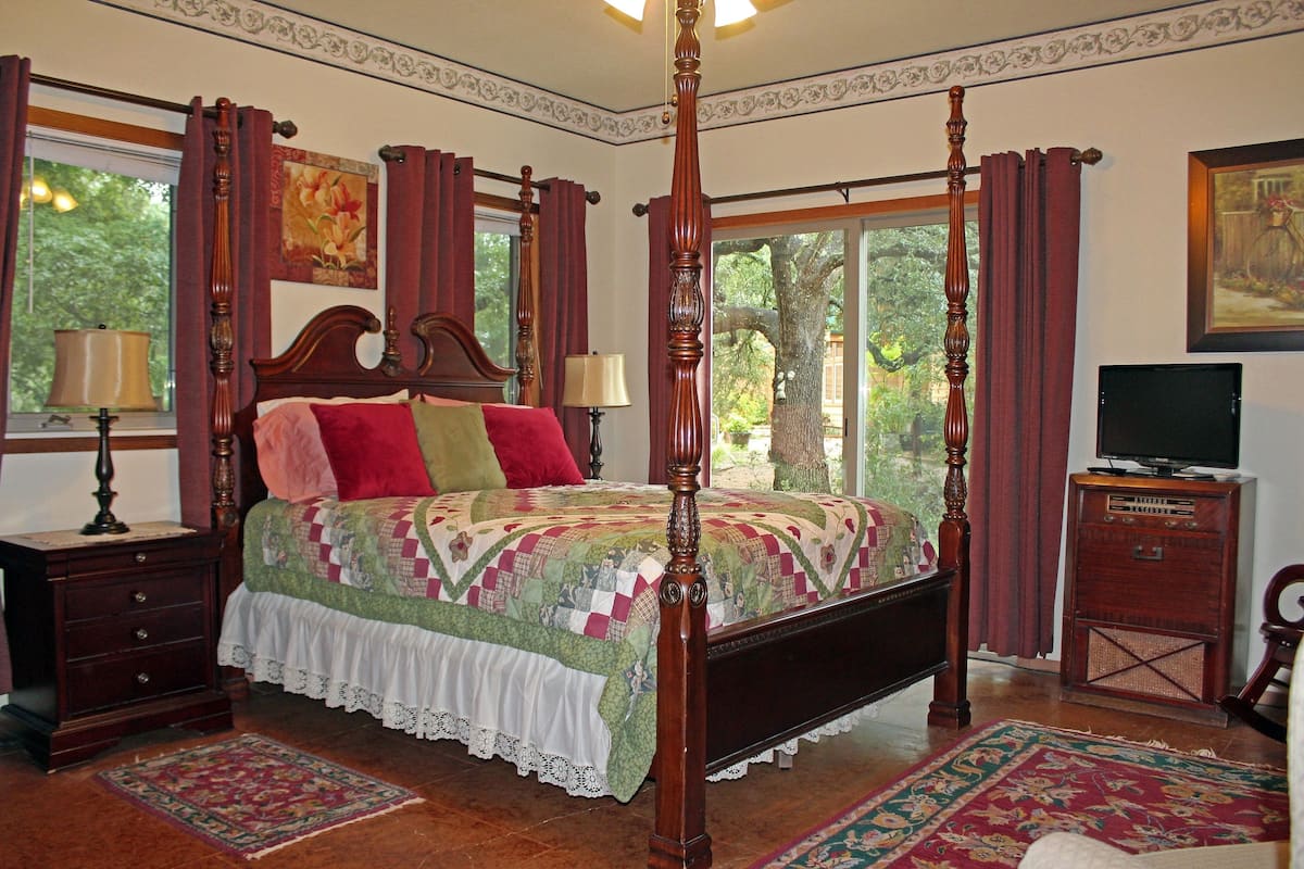 A four-poster bed with a quilt, a nightstand with a lamp, a dresser with a television, and a window with curtains.