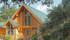 A wooden cabin with a green roof, large windows, and a front porch with potted plants. - Prow'd House Bed & Breakfast (Wimberley)