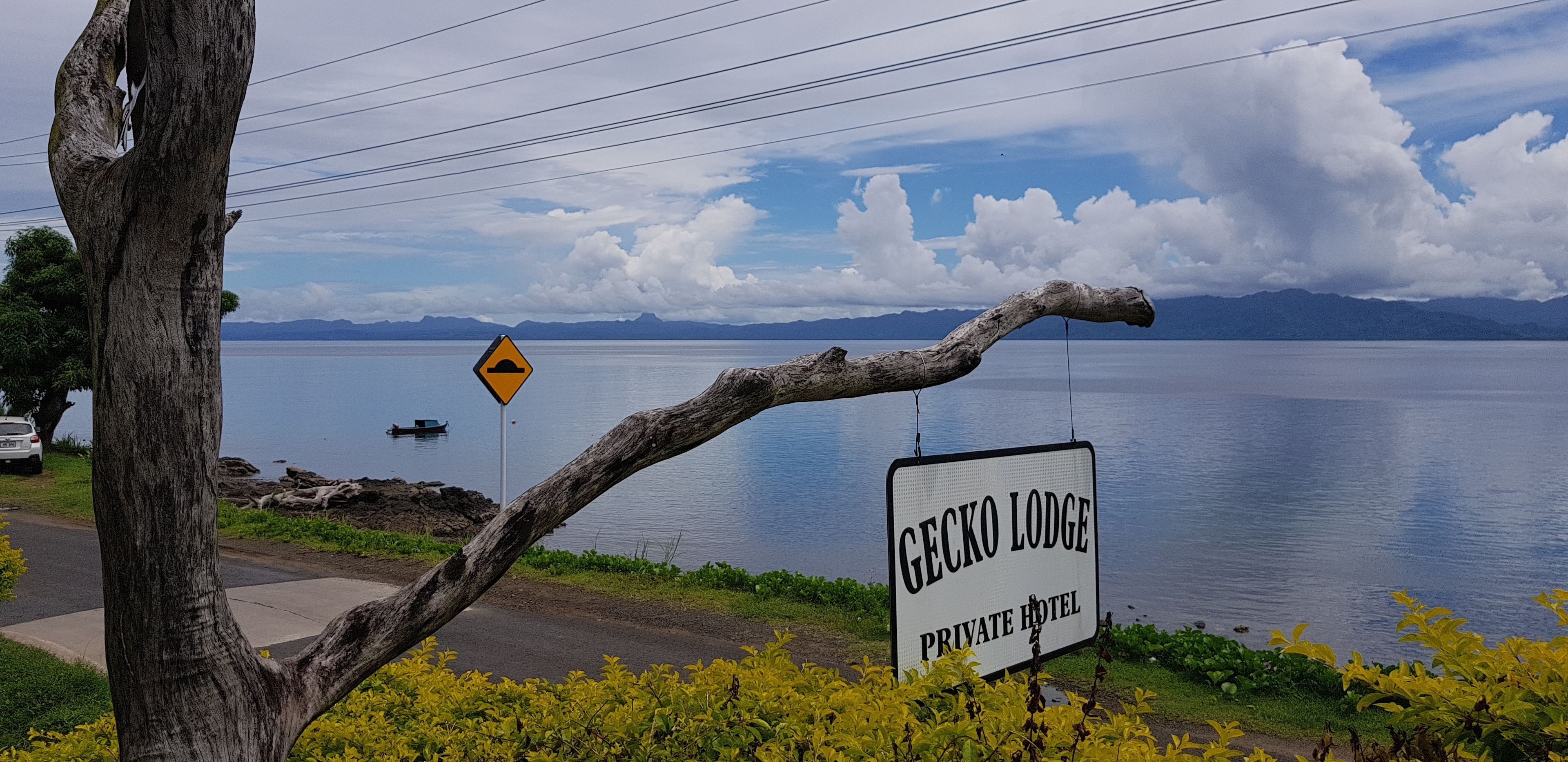 Cartel del Gecko Lodge Private Hotel junto a un lago, con un letrero de advertencia cerca.