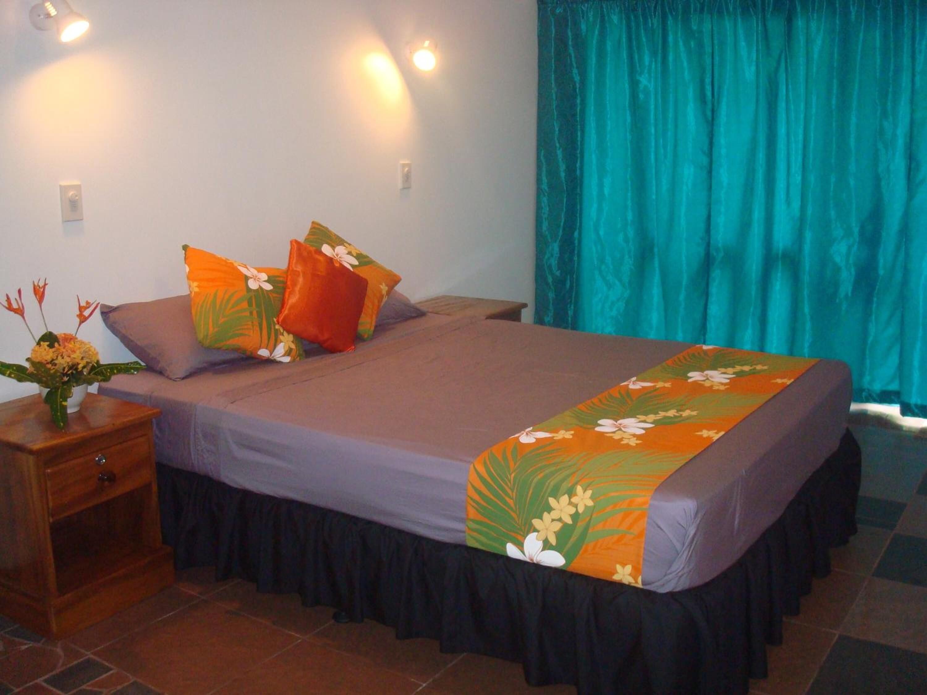 Una cama bien hecha con una colcha estampada con motivos florales, una mesita de noche con un jarrón de flores y una cortina color turquesa.