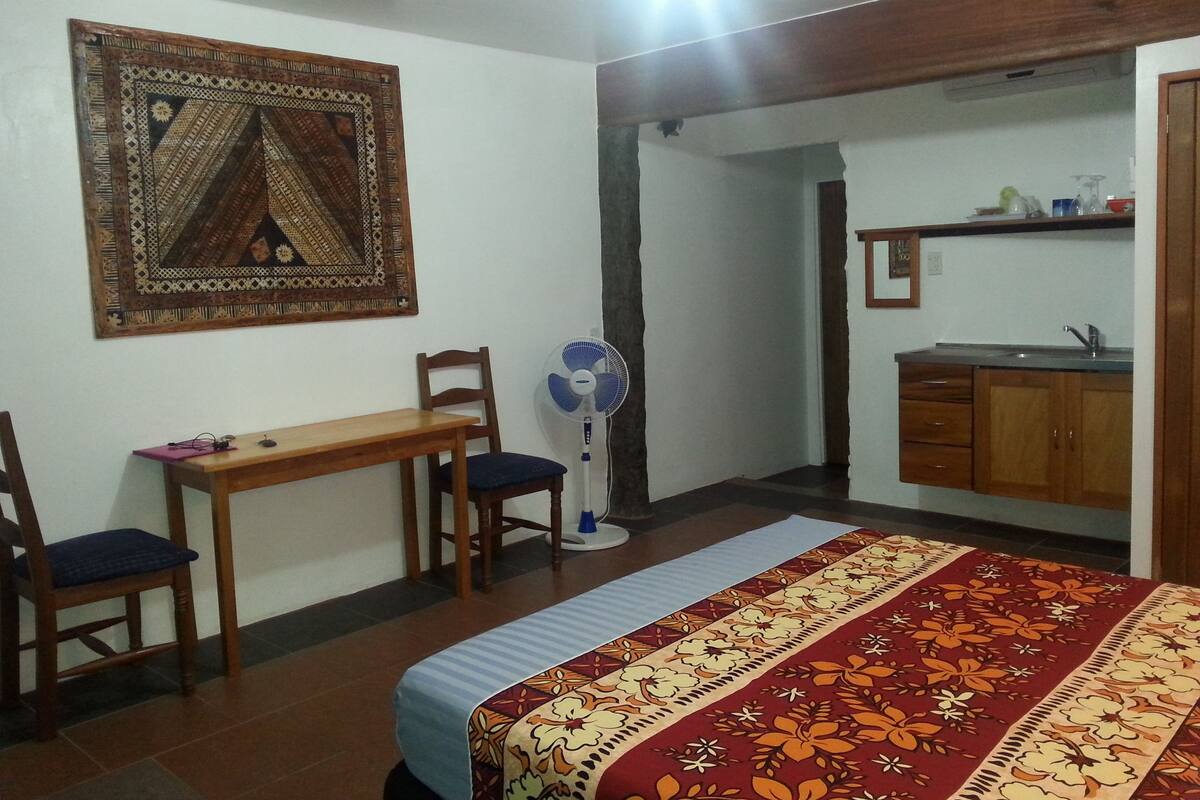 A bedroom with a bed, a desk, a chair, a standing fan, and a kitchen area with a sink and cabinets.
