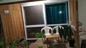 A room with a wooden wall, a large window with a curtain, a white chair, a small table, and a potted plant. - Gecko Lodge Fiji Private Hotel (Savusavu)