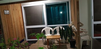 Um quarto com parede de madeira, uma janela grande com cortina, uma cadeira branca, uma mesinha e um vaso com planta.