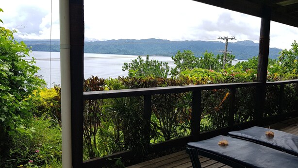 A covered outdoor area with a view of a lake and mountains. There are wooden tables and chairs. - Gecko Lodge Fiji Private Hotel (Savusavu)