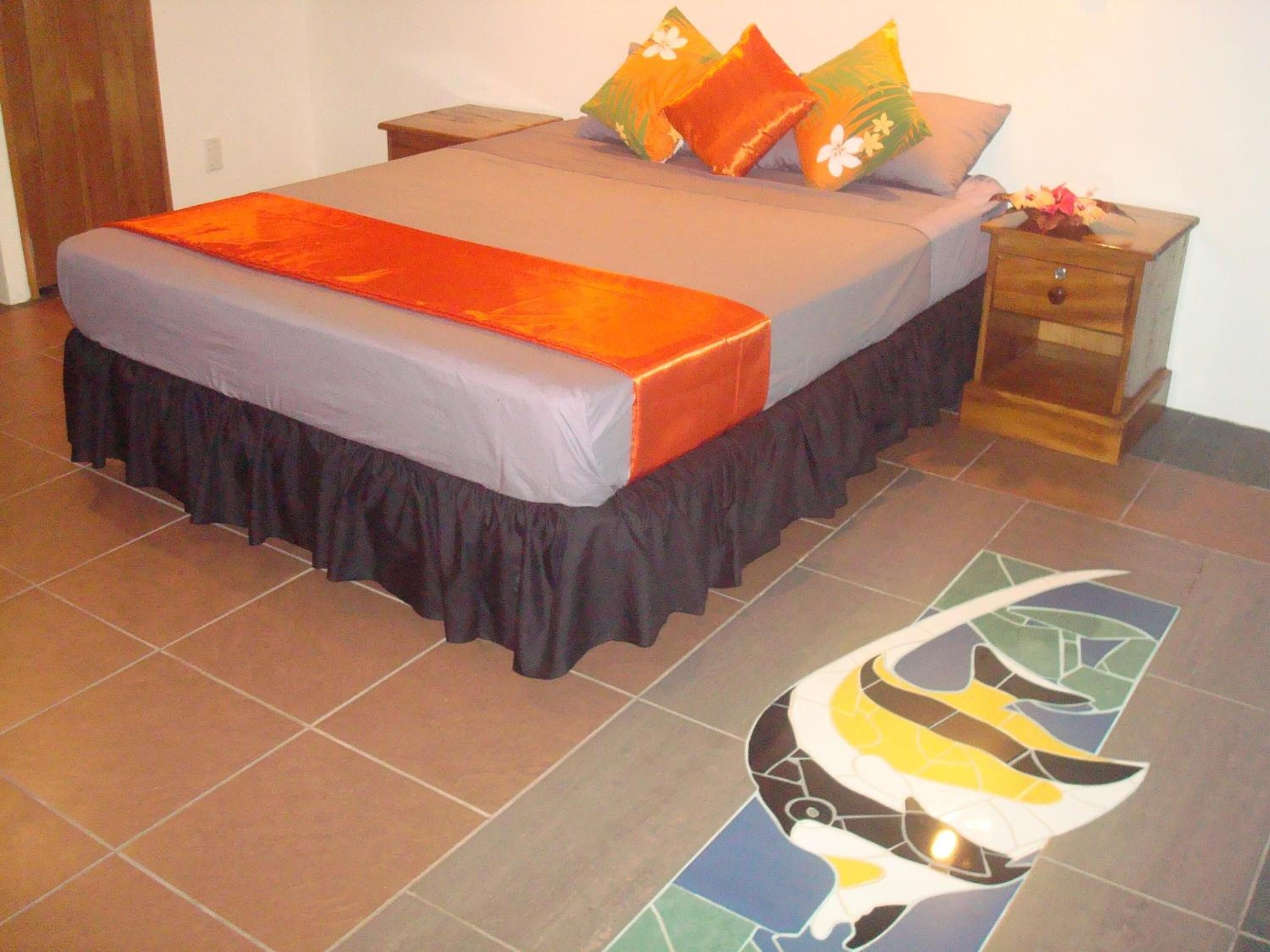 A bed with a grey and orange bedspread, a black skirted bed base, and a floral pillow arrangement. There is a wooden nightstand with a drawer and a small vase with flowers. The floor has a colorful mosaic design.