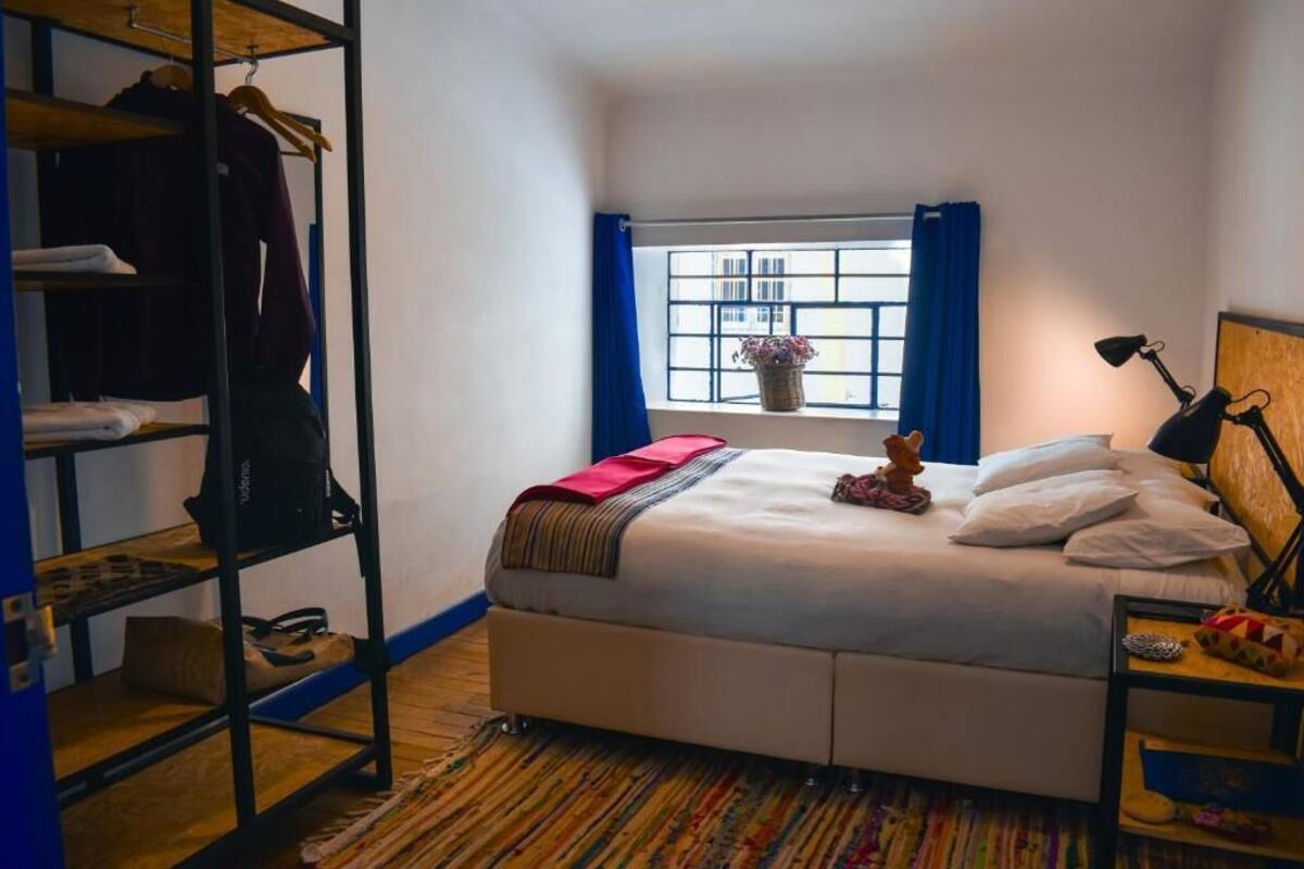 Ein Schlafzimmer mit Bett, Kleiderschrank, Fenster mit blauen Vorhängen, Schreibtisch mit Lampe und einer kleinen Pflanze auf der Fensterbank.