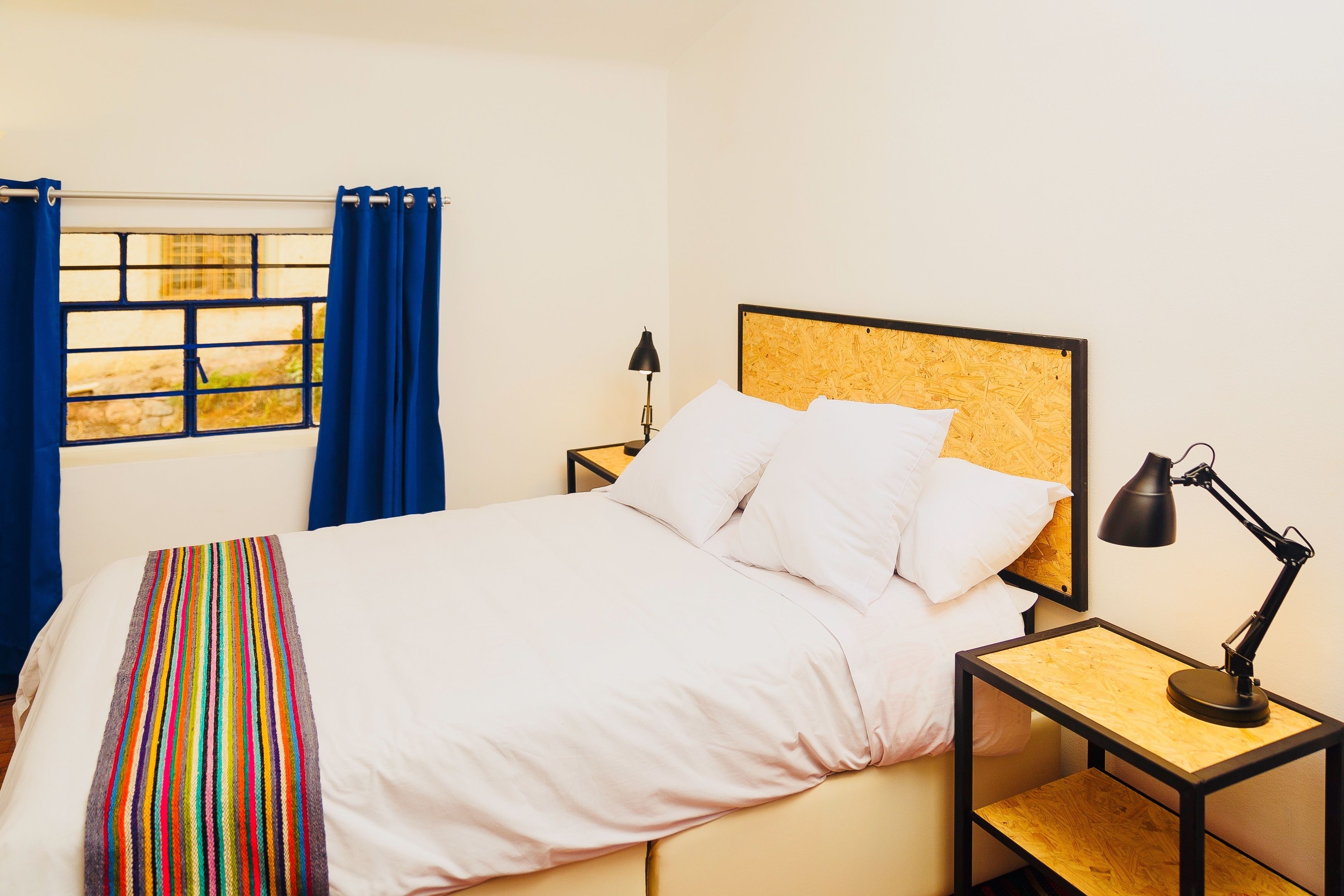Una cama bien hecha con sábanas blancas, cabecera de madera y mesita de noche con lámpara.