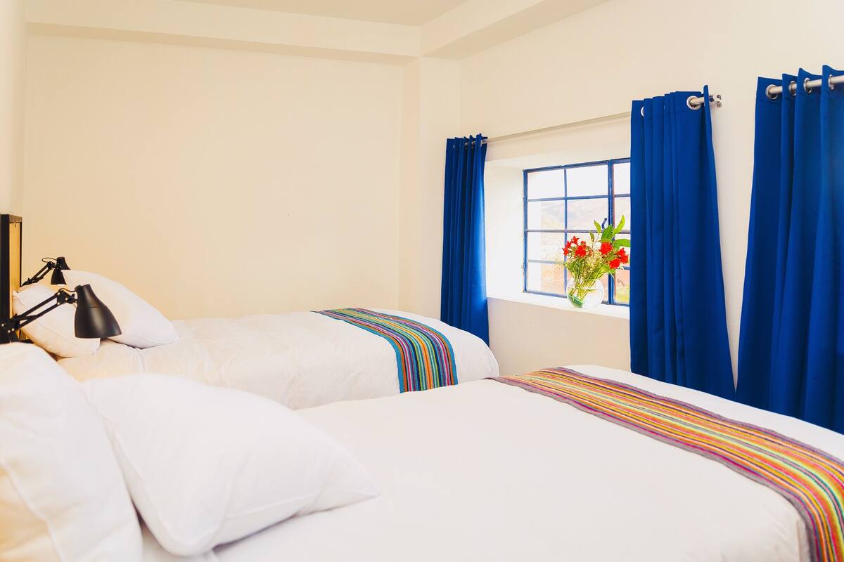 Ein Hotelzimmer mit zwei Betten, einem Fenster mit blauen Vorhängen, einer Nachttischlampe und einer Vase mit Blumen.