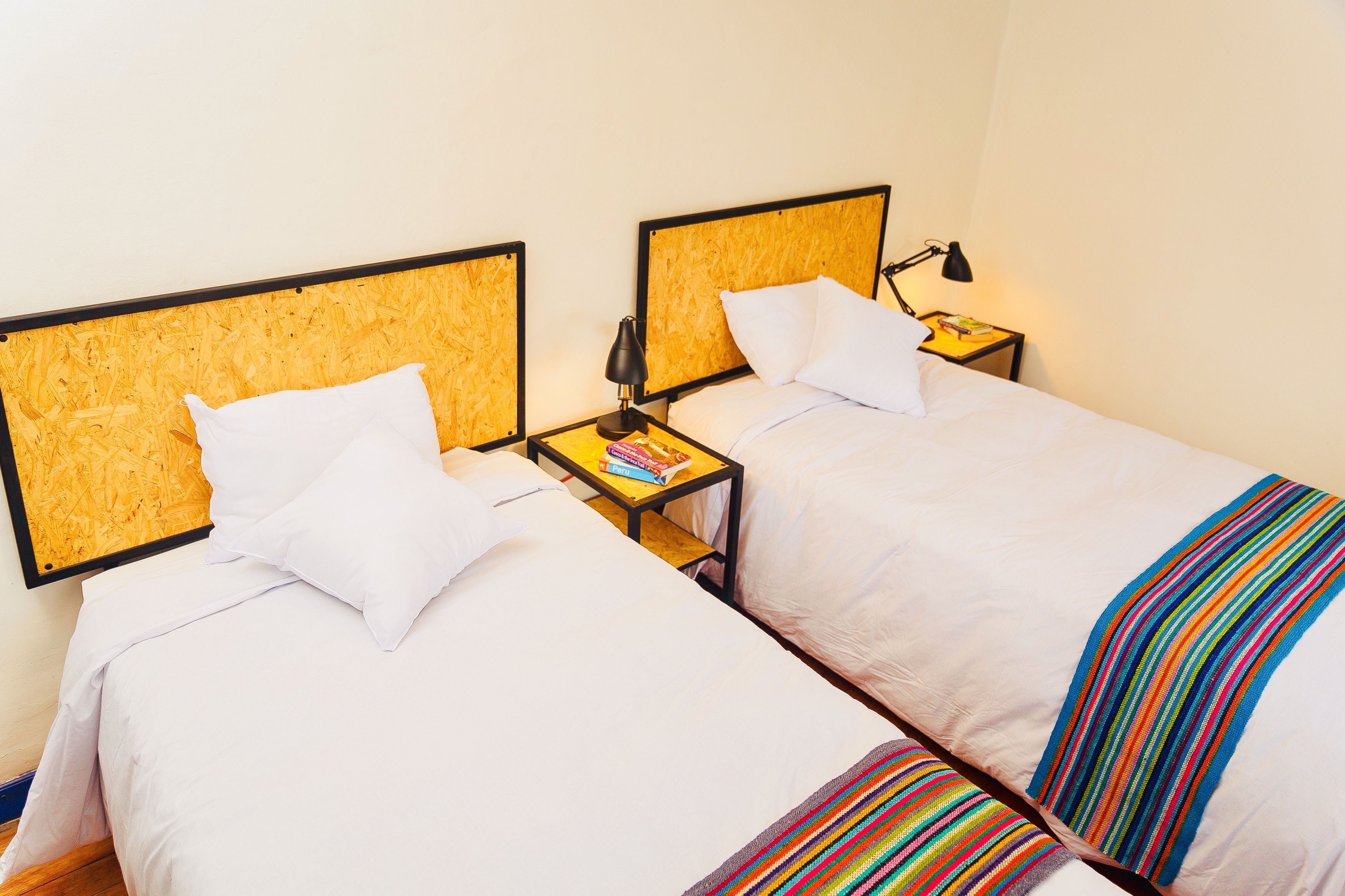 Una habitación de hotel con dos camas, cada una con cabecero de madera, ropa de cama blanca y una manta a rayas coloridas. Hay mesitas de noche con lámparas y libros.