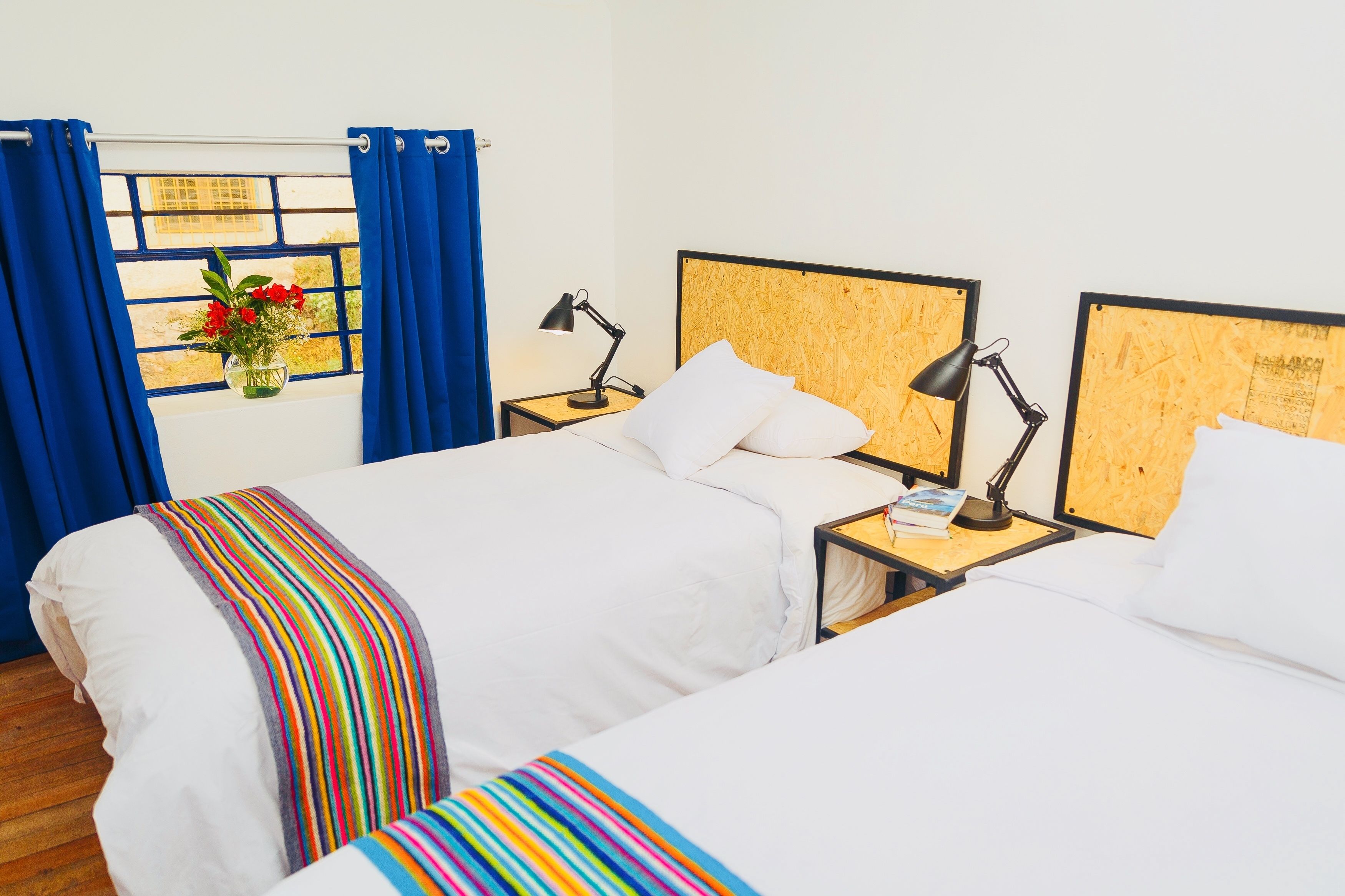 Una habitación de hotel con dos camas, una ventana con cortinas azules, una mesita de noche con una lámpara y un tablón de anuncios en la pared.