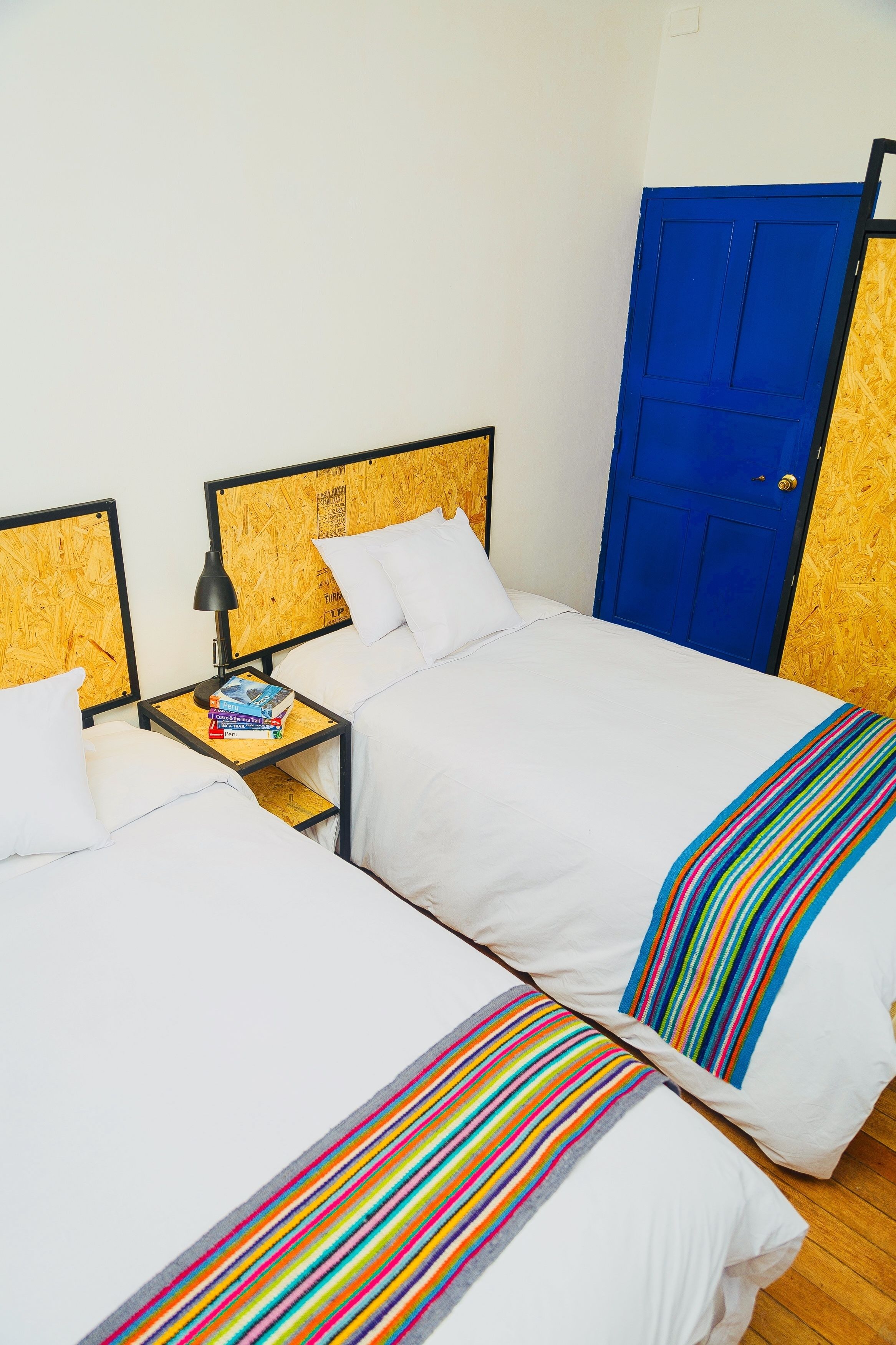A room with two beds, each with white bedding and colorful striped pillows. There is a nightstand with a lamp and books between the beds. A blue door is visible in the background.