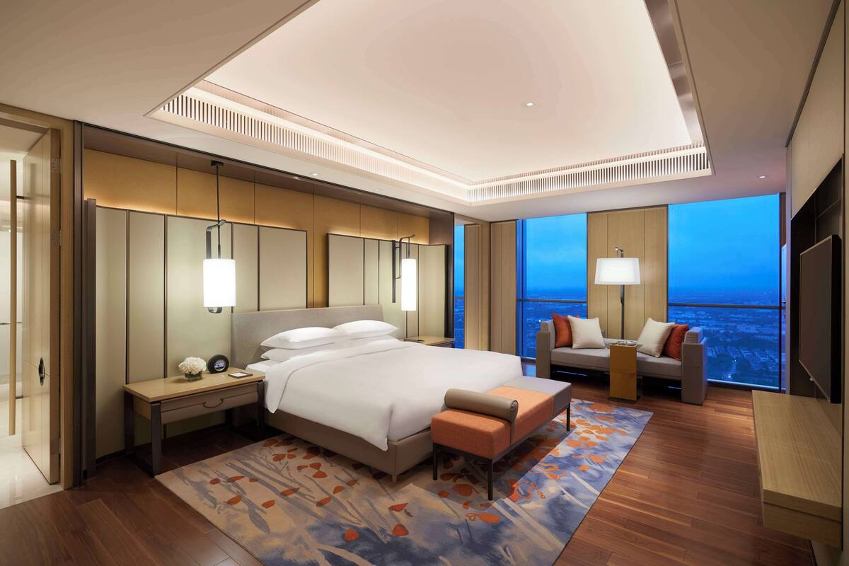 A modern hotel room with a large bed, a sitting area with a sofa and armchair, a desk, and a view of the city at dusk.