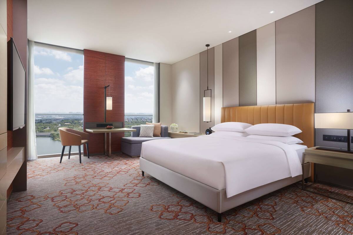 A modern hotel room with a large bed, a desk, a chair, and a view of the cityscape.