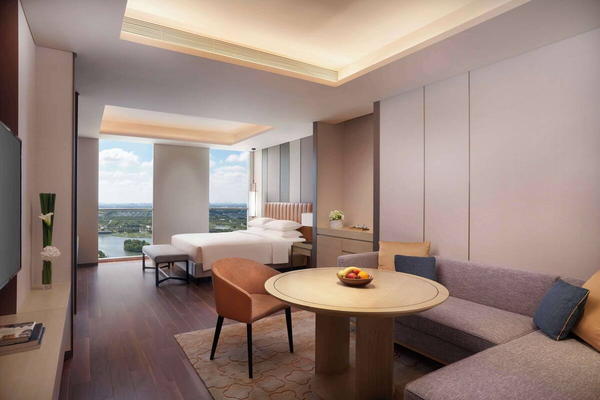 A modern hotel room with a large bed, a sofa, a round table with fruit, and a view of the city.