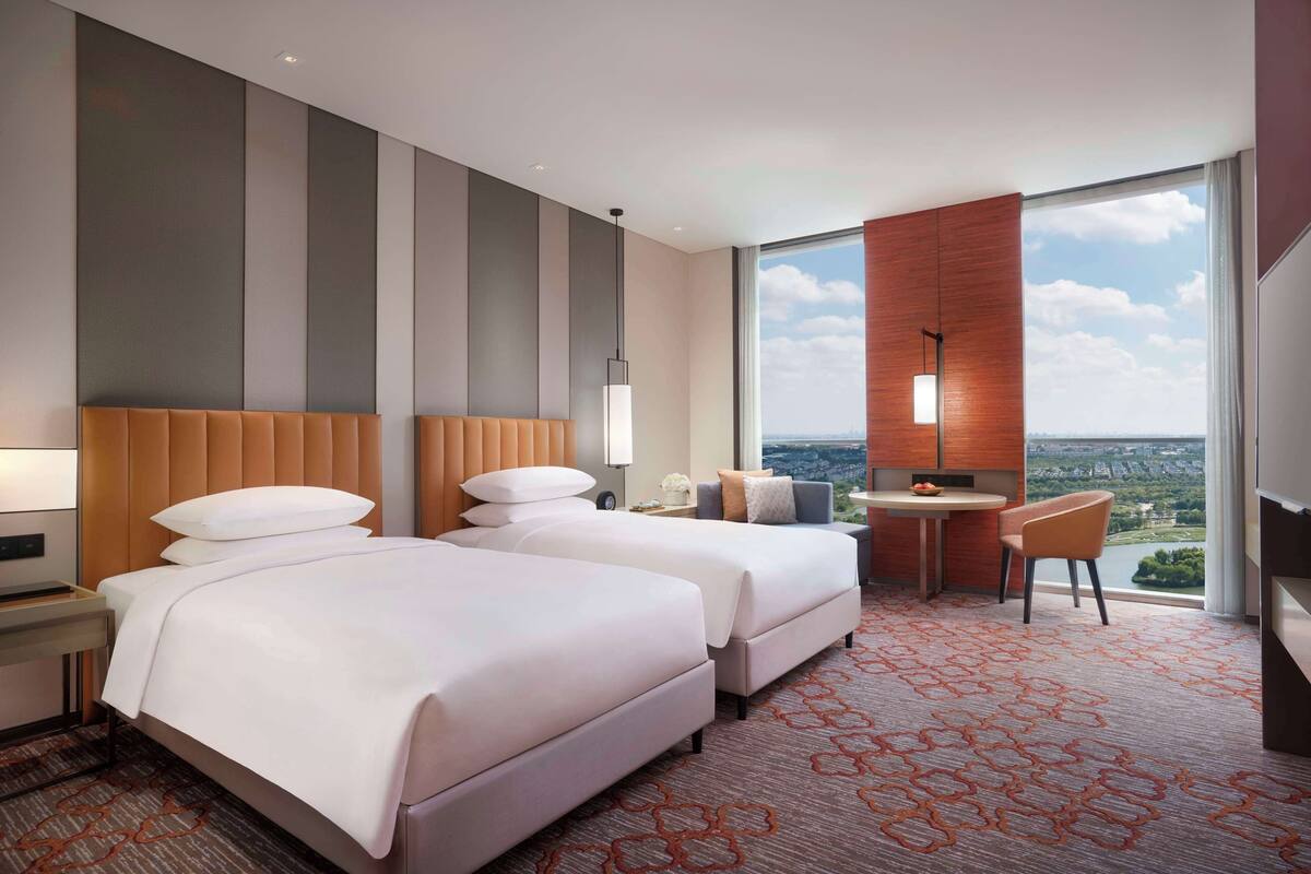 A hotel room with two beds, a desk, a chair, and a large window offering a city view.