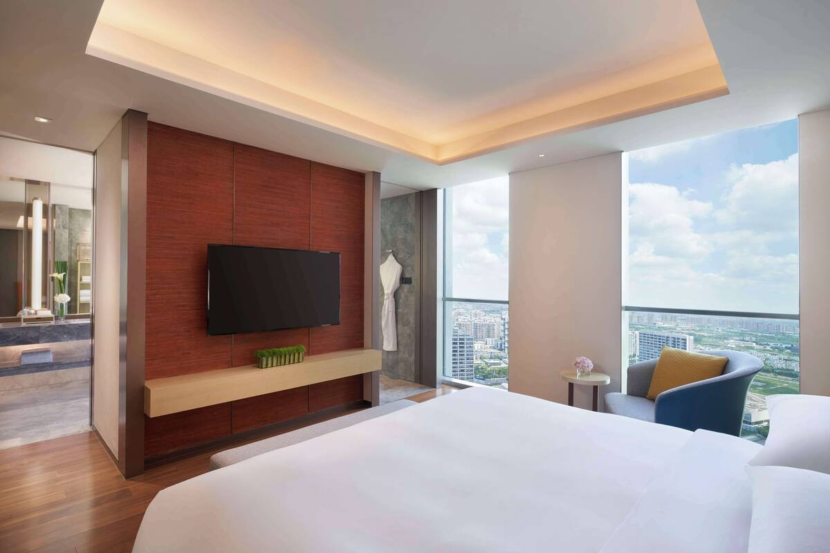 A modern hotel room with a large bed, a flat-screen TV mounted on a wooden panel, a small table with a vase, and a chair with a yellow cushion.