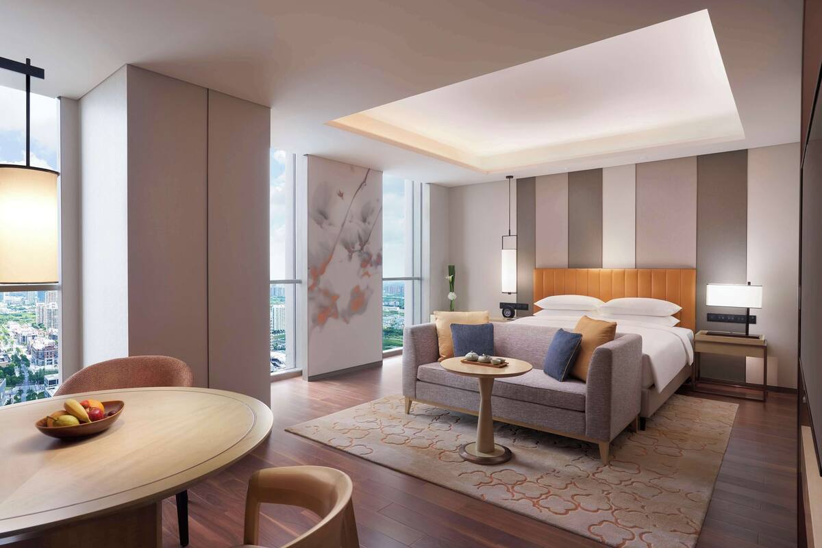 A modern hotel room with a bed, a sofa, a dining table, and a view of the city.