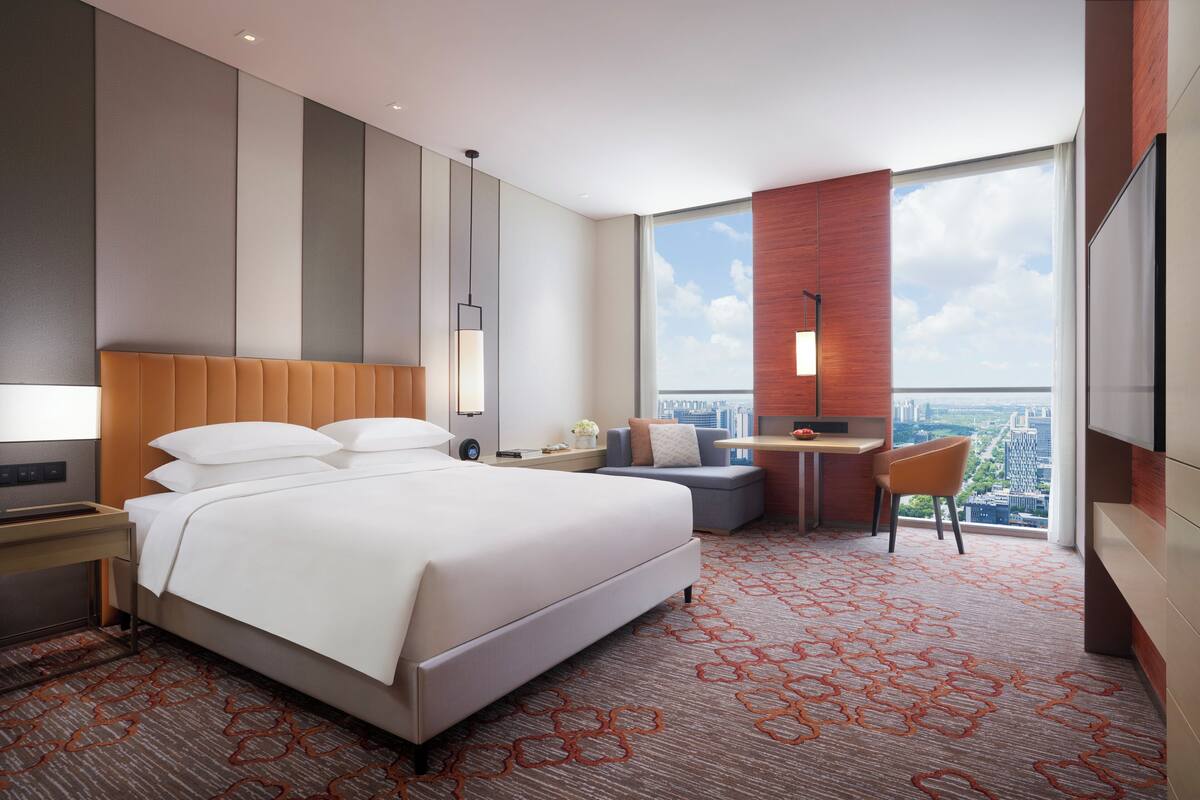 A hotel room with a large bed, a desk, a chair, and a TV. There is a view of the city from the window.