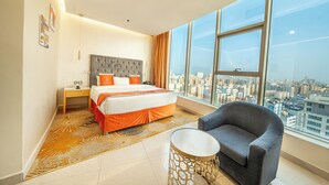 Habitación de hotel moderna con una cama grande, un sillón gris, una mesita redonda y vistas a la ciudad a través de amplias ventanas.
