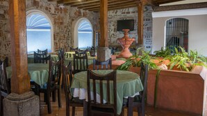Un comedor rústico con mesas y sillas de madera, una maceta de terracota grande y vistas a través de un ventanal arqueado.