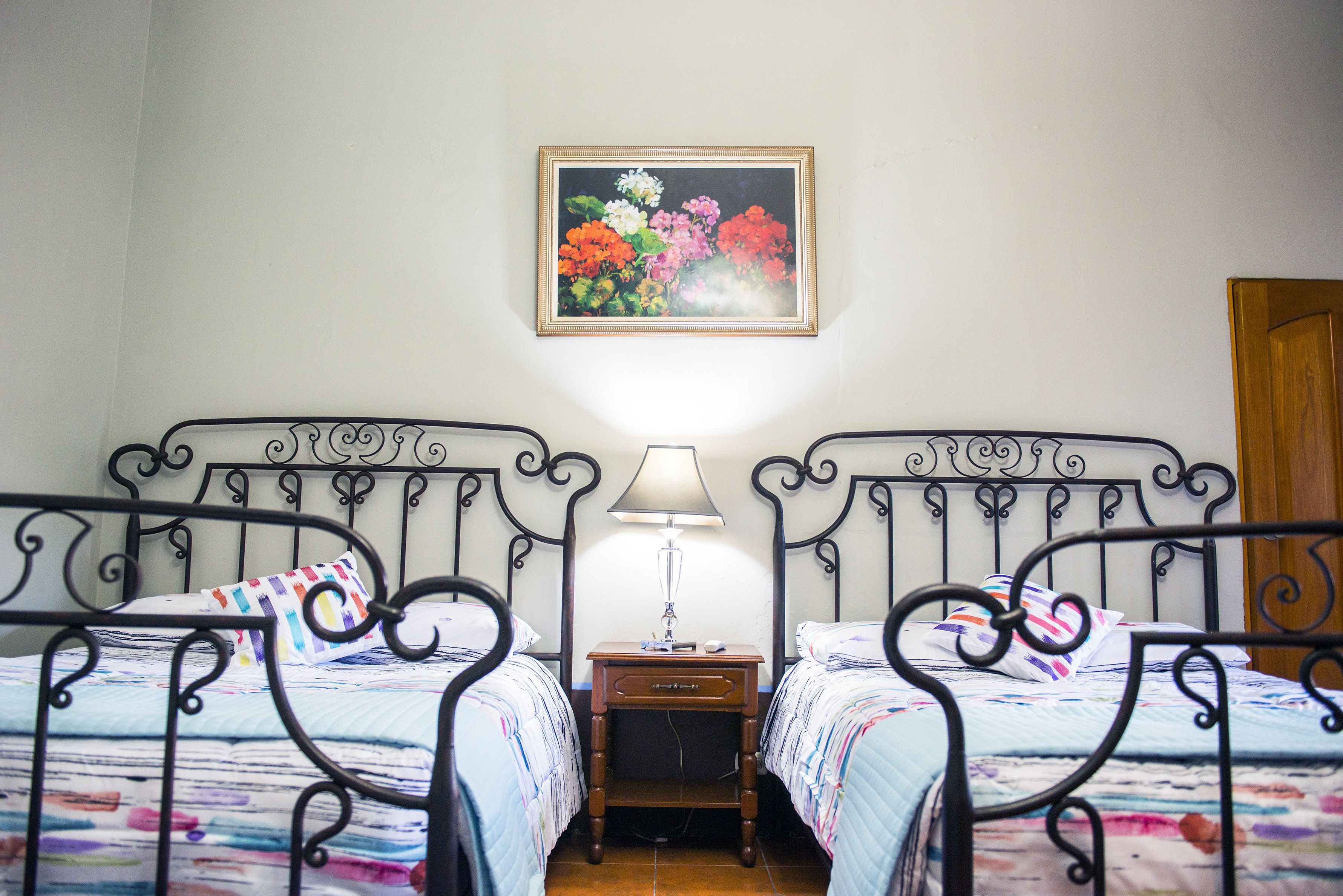 A room with two beds, a nightstand with a lamp, and a floral painting on the wall.
