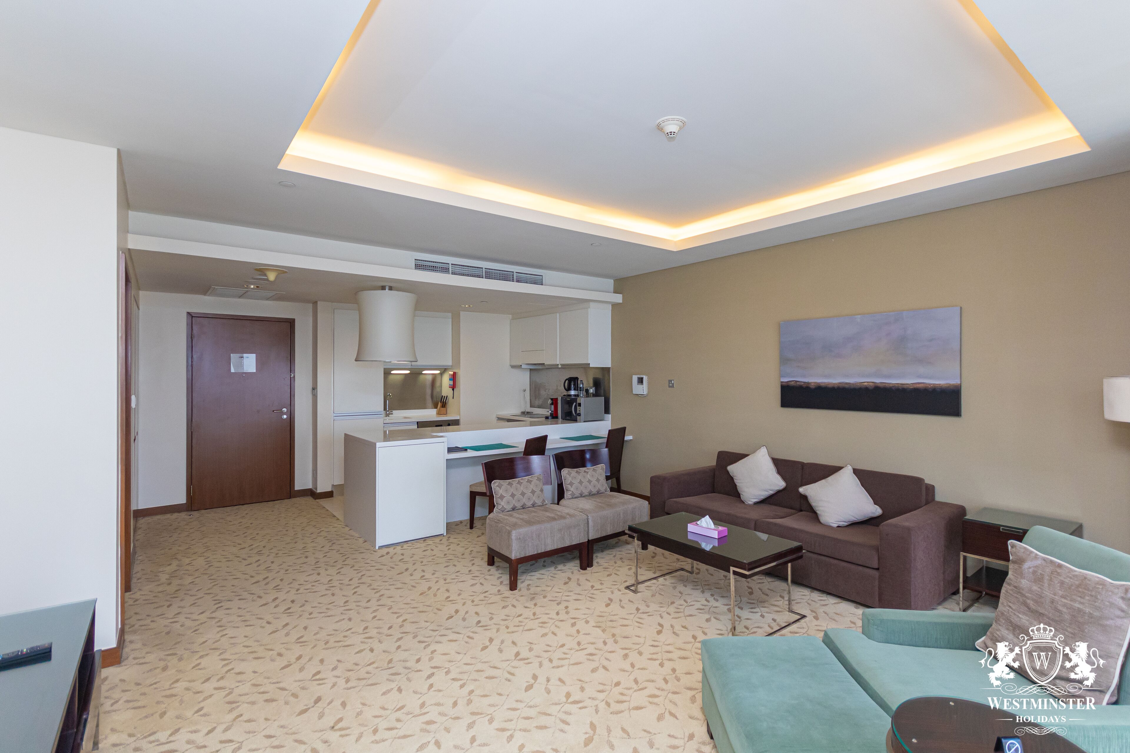 A modern living room with a sofa, armchairs, and a coffee table. There is a kitchenette in the background with a microwave and a coffee maker.