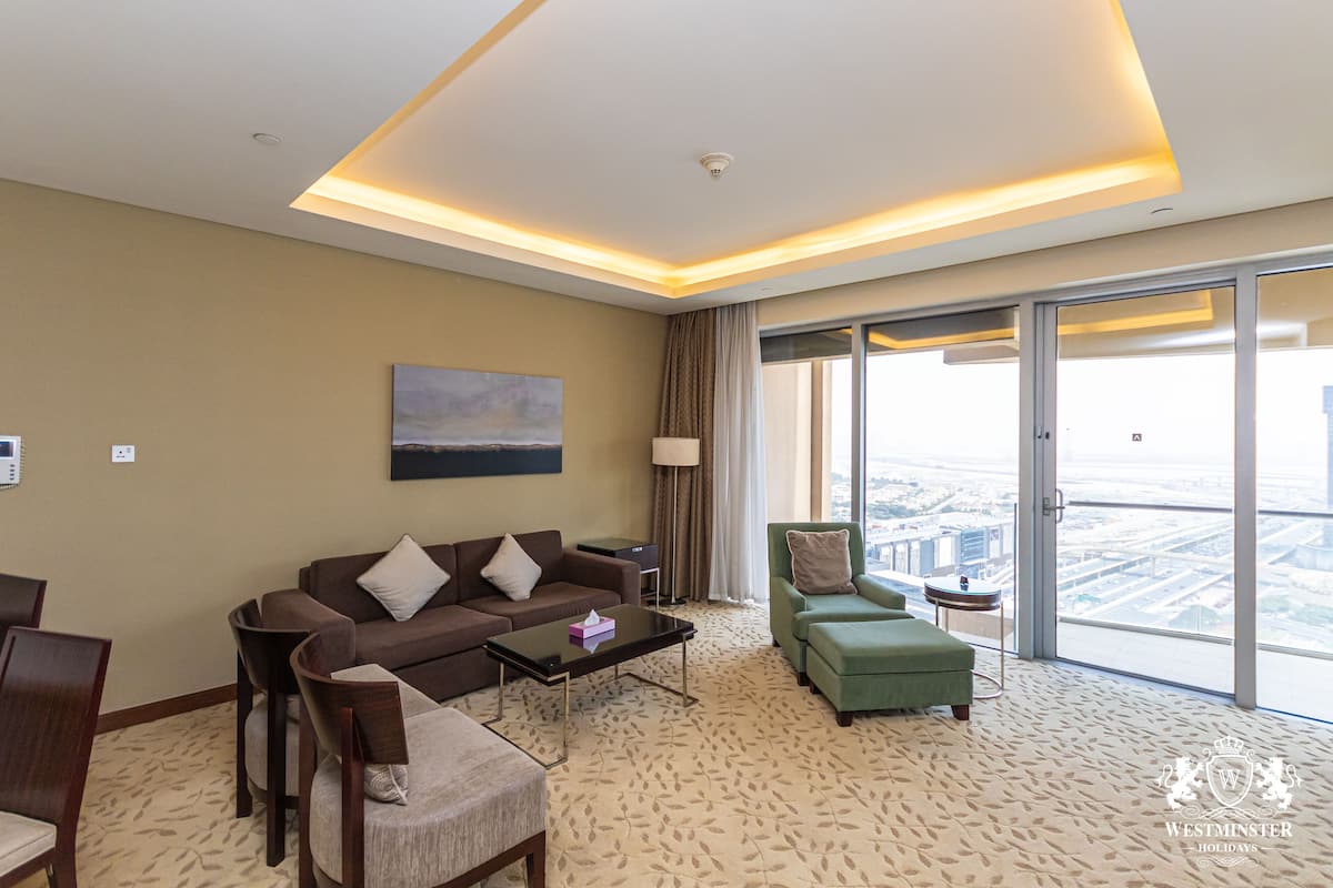 A modern hotel room with a large window offering a city view, a sofa, a green armchair, and a coffee table.