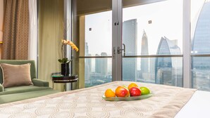 Habitación de hotel con cama, un sillón verde, una mesita con un florero y un frutero sobre la cama. La habitación tiene ventanas amplias con vistas a la ciudad.