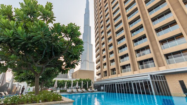 Una piscina con sillones de descanso y sombrillas, rodeada de follaje, con un edificio alto al fondo.