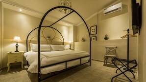 Een hemelbed met een kroonluchter, nachtkastjes met lampen, een stoel en een wandrek.